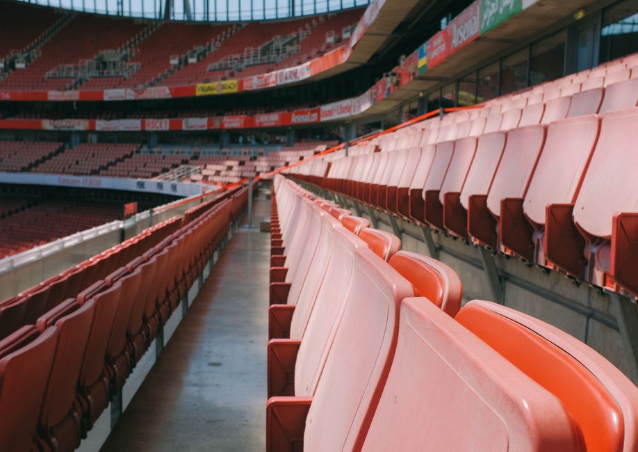 Empty stadium seating with rows of pink and red plastic chairs arranged along a concrete walkway. The seats are clean and well-maintained, with no visible dust or debris. Bright lighting highlights th