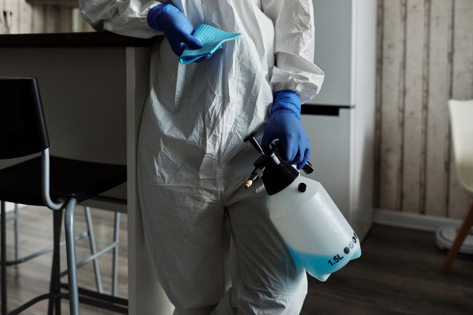 A close-up of a professional cleaner wearing a white protective suit and blue gloves, holding a black spray gun with a white 1.5-liter container filled with cleaning solution. The cleaner is spraying 